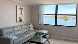 A modern living room with a sectional sofa, a glass coffee table, and a large window offering a view of a body of water and palm trees. - Charming condo in fantastic St. Petersburg Beach, with waterfront, sunset views (St. Petersburg)