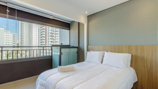 A modern hotel room with a large bed, a view of tall buildings, and a balcony with a railing. - Complete Studio With Self Check-in Cob0710 (São Paulo)