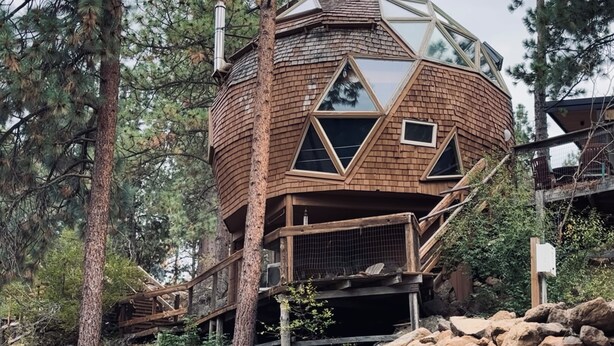 A geodesic dome structure with a wooden exterior, surrounded by trees and rocks.