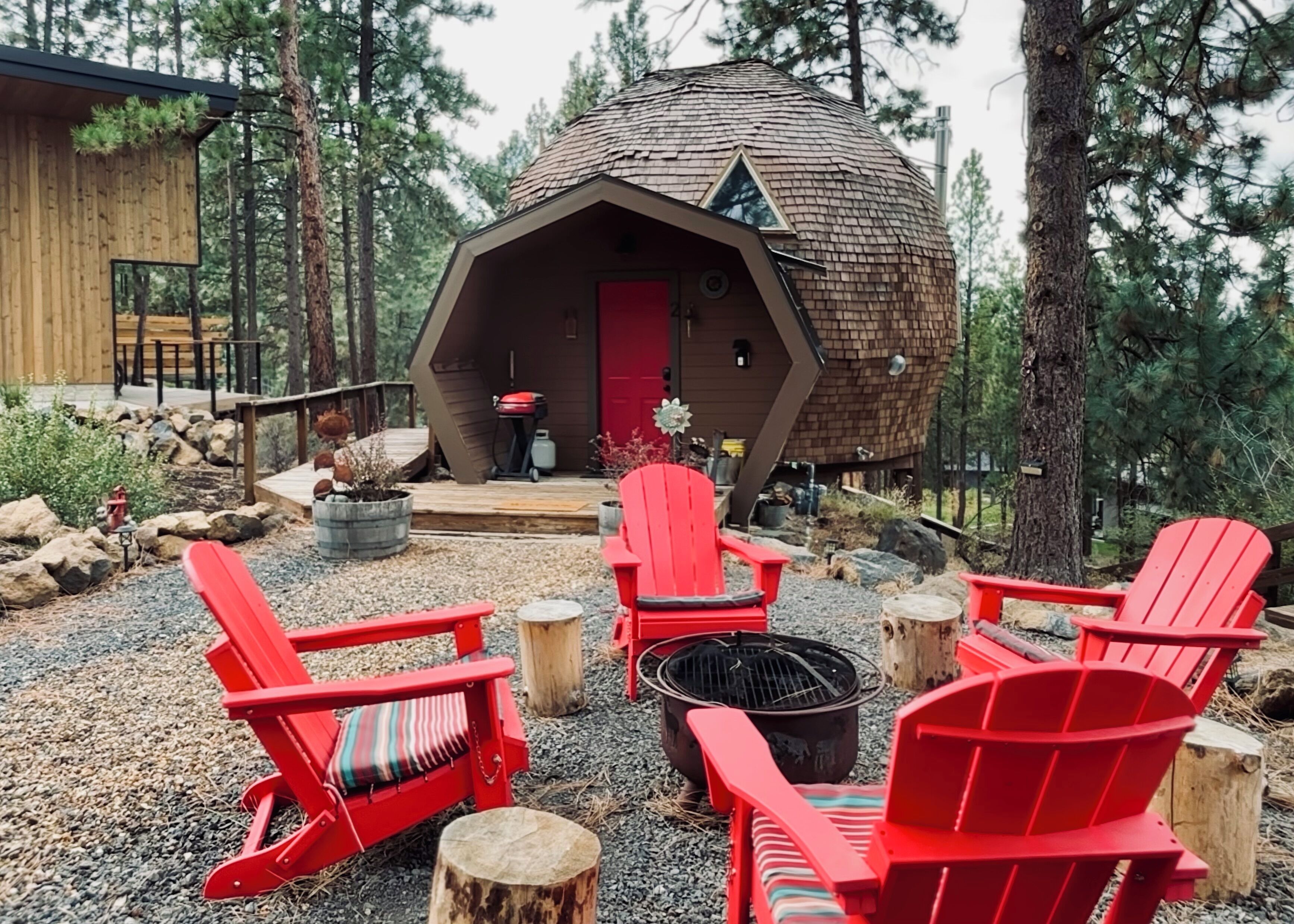 A cozy cabin with a conical roof, surrounded by red Adirondack chairs, a fire pit, and a deck leading to the entrance.