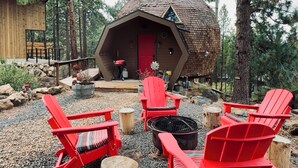 A cozy cabin with a conical roof, surrounded by red Adirondack chairs, a fire pit, and a deck leading to the entrance.