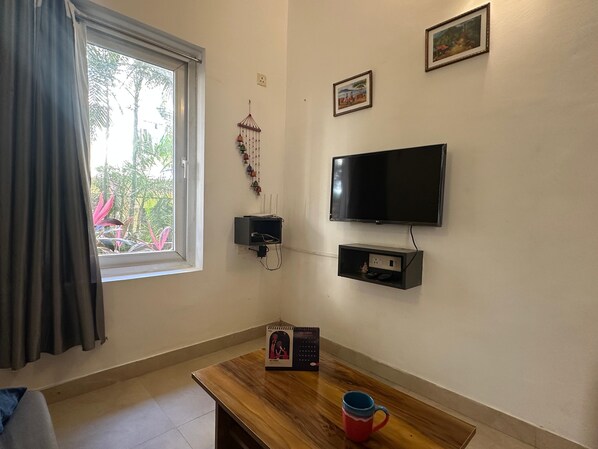 Una habitación con una mesa de madera, un televisor montado en la pared, una ventana con cortinas y un estante pequeño con un objeto decorativo.