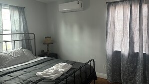Quarto com cama, mesa de cabeceira, luminária, janela com cortinas e ar-condicionado.