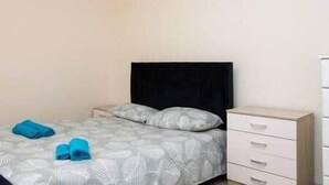 Una cama individual con cabecera negra, ropa de cama gris estampada y dos toallas azules a un lado.