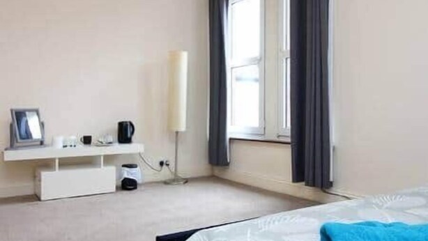 Un dormitorio con cama, mesita de noche, ventana con cortinas y estante en la pared con un marco de fotos y una taza.