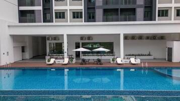 Piscina com espreguiçadeiras e guarda-sóis, área de estar coberta e um edifício com varandas.