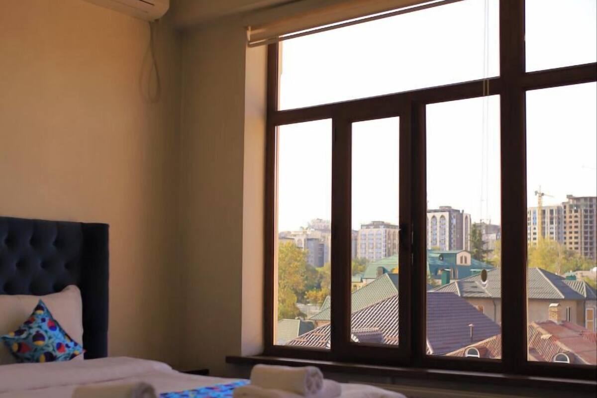 Ein Hotelzimmer mit einem großen Bett, Blick auf die Stadt durch ein Fenster und einer modernen Deckenleuchte.