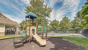 Une aire de jeux avec un toboggan, des éléments d’escalade et un espace abrité, le tout entouré d’une clôture et d’arbres.