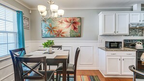 Un coin repas comprenant une table, des chaises, un lustre, ainsi qu’une cuisine avec des meubles blancs et des appareils électroménagers.