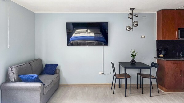 Una sala de estar moderna con un sofá gris, una pequeña mesa de comedor con dos sillas, un televisor montado en la pared y una zona de cocina con armarios de madera y una encimera negra.