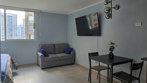 A modern living room with a grey sofa, a dining table with chairs, a flat-screen TV mounted on the wall, and a window with a view of tall buildings. - Modern studio close to Barrio Italia (Santiago)