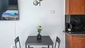 A compact living space with a dining table, two chairs, a small potted plant, a modern light fixture, a flat-screen TV mounted on the wall, and a wooden cabinet. - Modern studio close to Barrio Italia (Santiago)