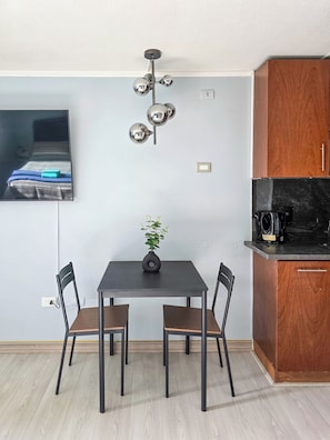 A compact living space with a dining table, two chairs, a small potted plant, a modern light fixture, a flat-screen TV mounted on the wall, and a wooden cabinet. - Modern studio close to Barrio Italia (Santiago)