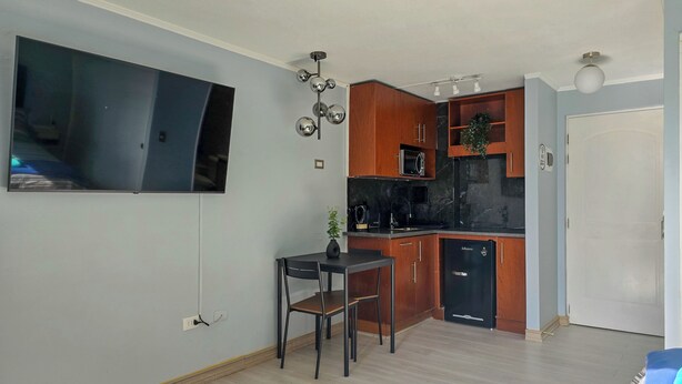 A compact kitchen with wooden cabinets, a black refrigerator, and a microwave. There's a small dining table with two chairs. A flat-screen TV is mounted on the wall. - Modern studio close to Barrio Italia (Santiago)