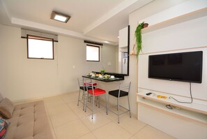 A modern living room with a sofa, a dining table with chairs, a flat-screen TV mounted on the wall, and a small shelf with decorative items. - Taiamã - Rua das Gaivotas - Ingleses (Florianópolis)