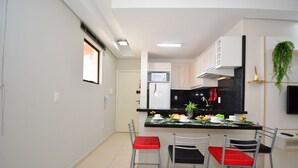 A modern kitchen with a central island, white cabinets, and a black countertop. There are two chairs with red cushions and a TV mounted on the wall. - Taiamã - Rua das Gaivotas - Ingleses (Florianópolis)