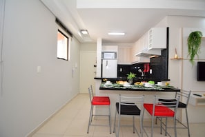Una cocina moderna con una isla central, armarios blancos y encimera negra. Hay dos sillas con cojines rojos y un televisor montado en la pared.