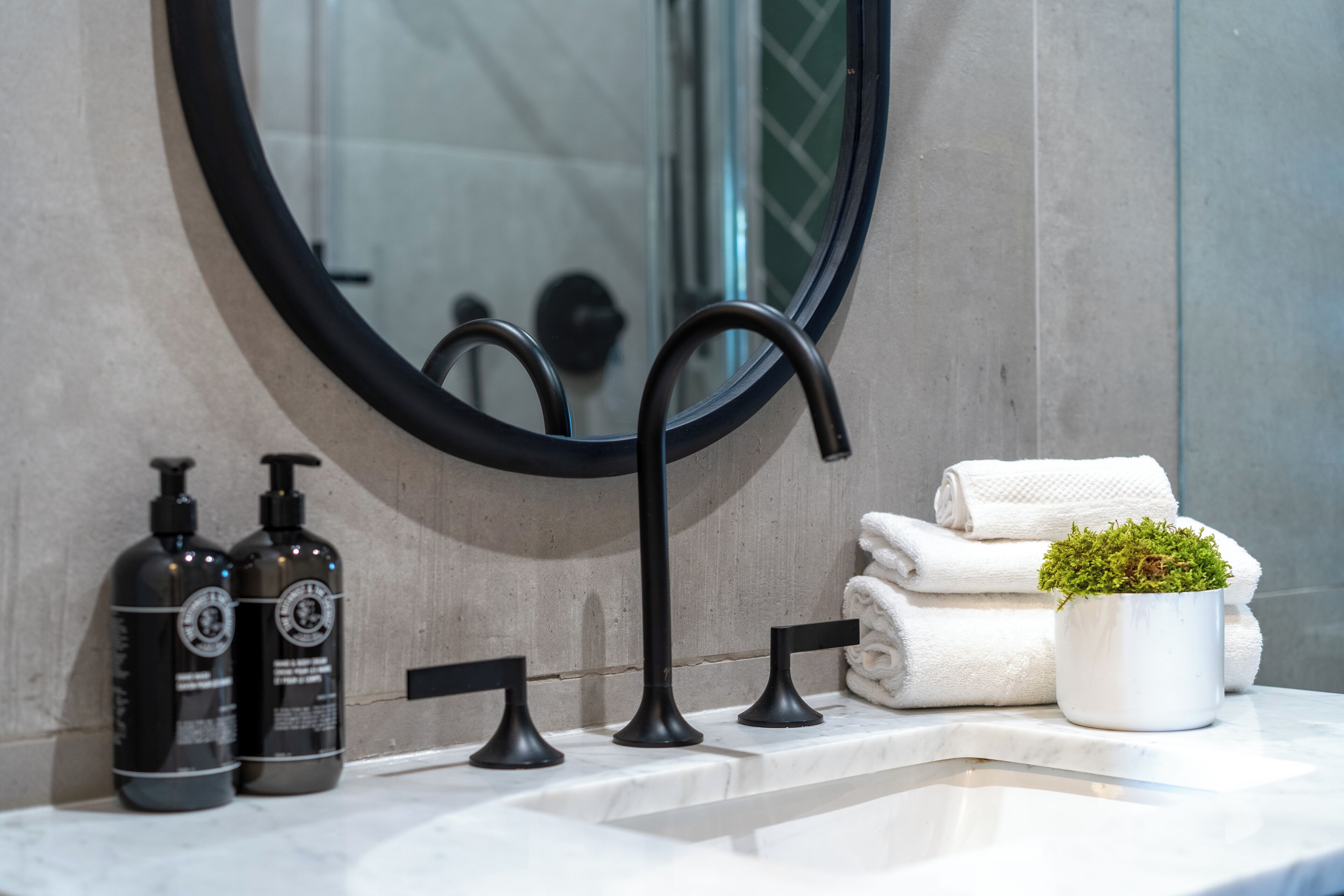 A bathroom with a round mirror, a black faucet, two bottles of shampoo, a potted plant, and stacked towels.