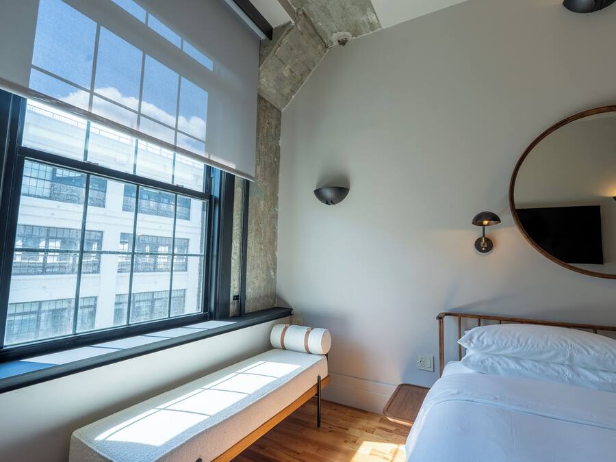 Un dormitorio con cama, un banco junto a la ventana, un espejo redondo e iluminación en la pared.