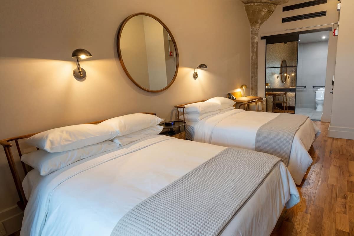 Hotellihuone, jossa on kaksi sänkyä, pyöreä peili, seinään kiinnitetyt lamput ja pieni pöytä.