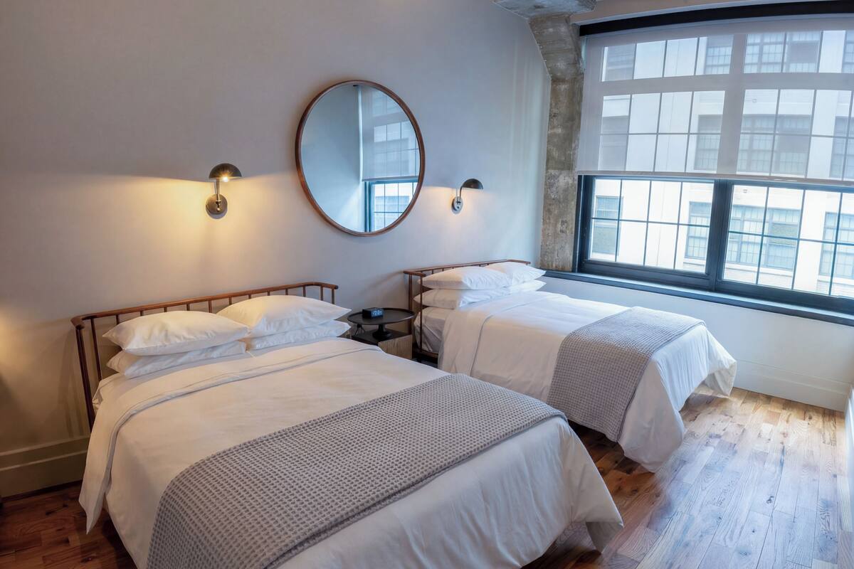 Hotellihuone, jossa on kaksi sänkyä, suuri ikkuna ja pyöreä peili seinällä.