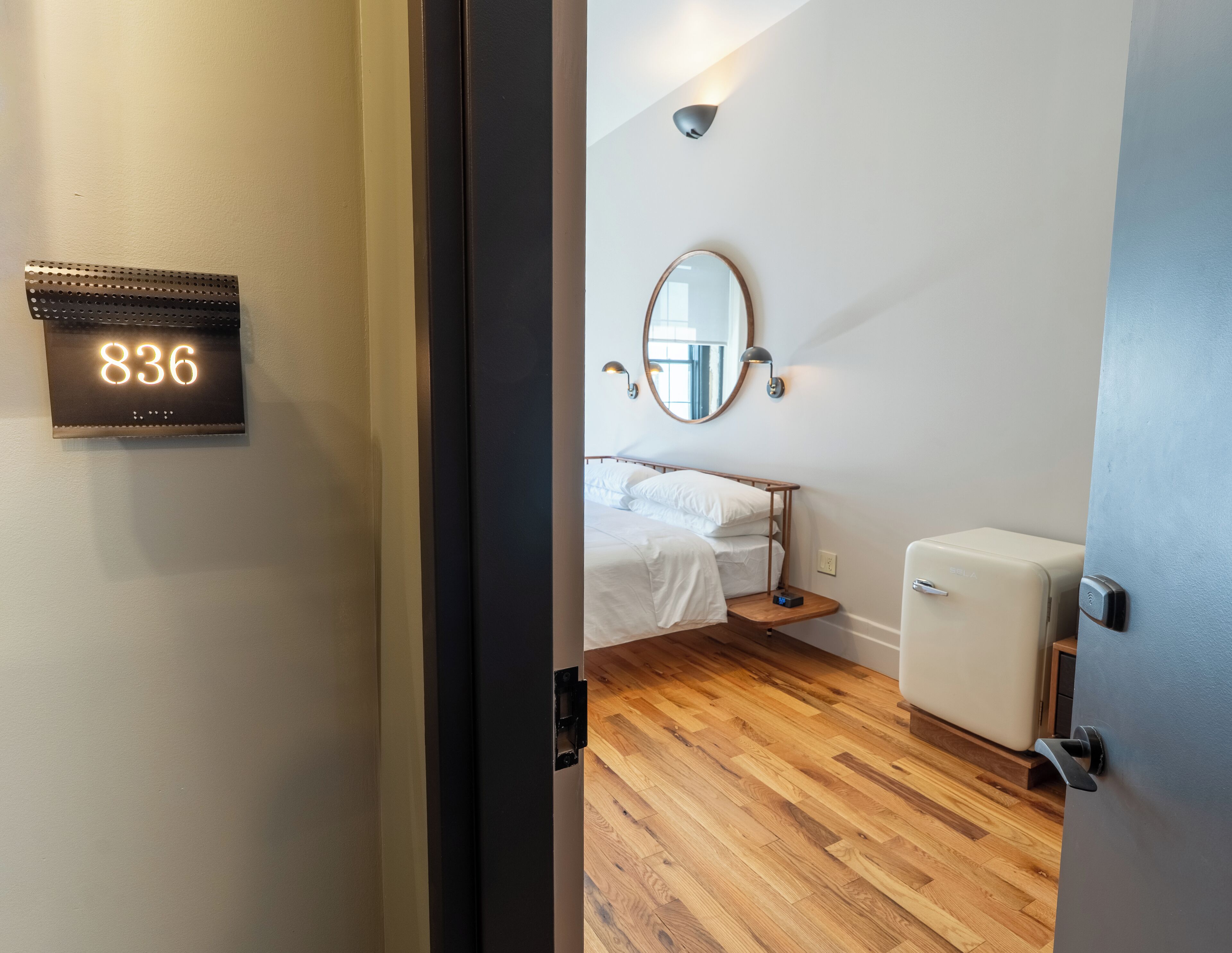 Habitación de hotel con cama, mesita de noche, espejo, luz de pared y puerta con un número.