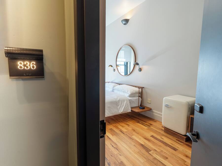 Habitación de hotel con cama, mesita de noche, espejo, luz de pared y puerta con un número.