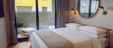 A hotel room with a large bed, a round mirror, a bedside table, and a view of a building through a glass door.
