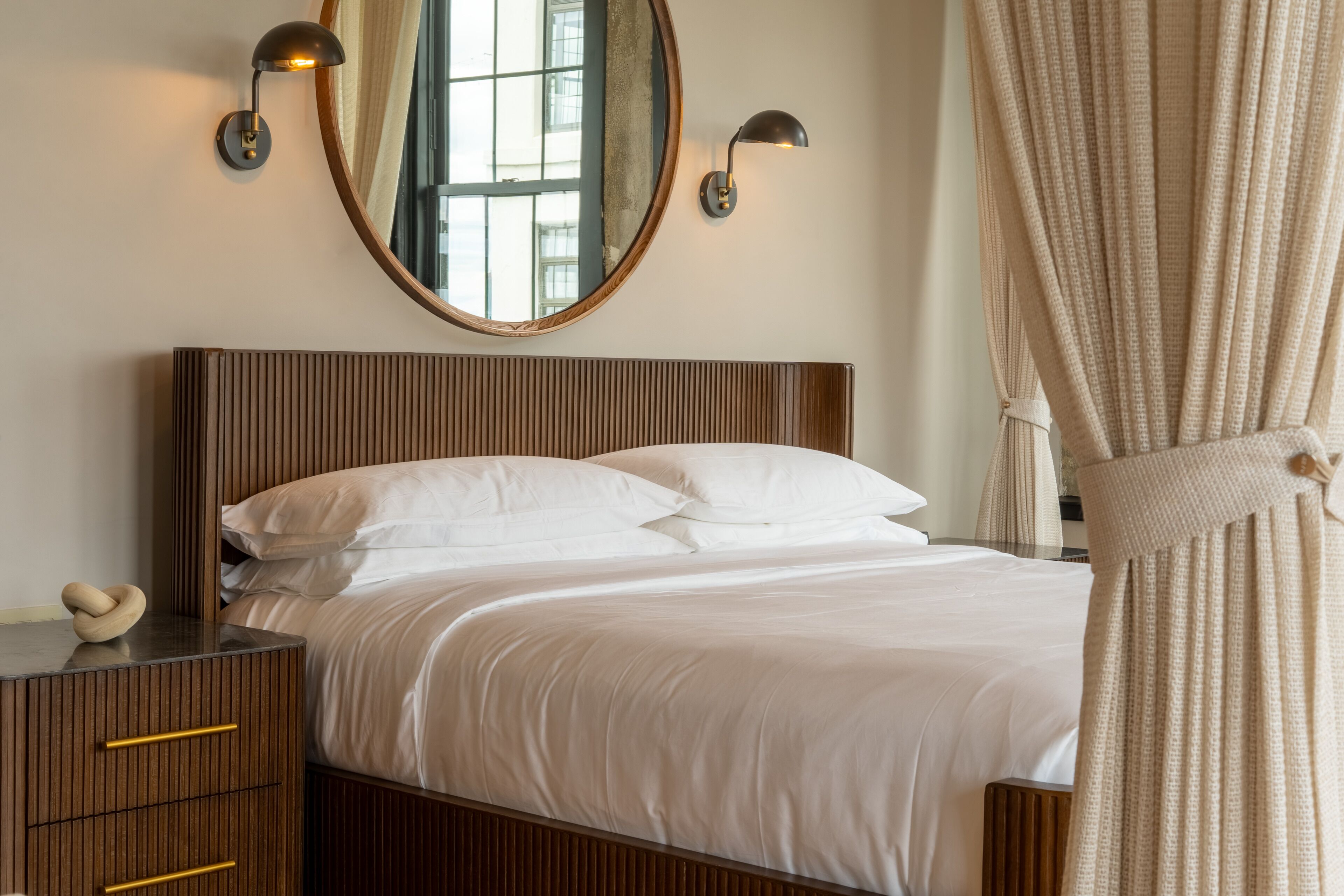 A neatly made bed with white linens, a wooden headboard, and two wall-mounted lamps.