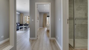 A hallway with a glass shower door, a sofa, and a dining table in the background.