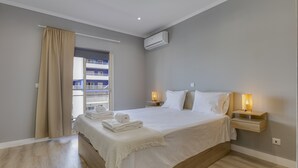 A bedroom with a large bed, a bedside table with a lamp, an air conditioning unit, and a window with curtains.
