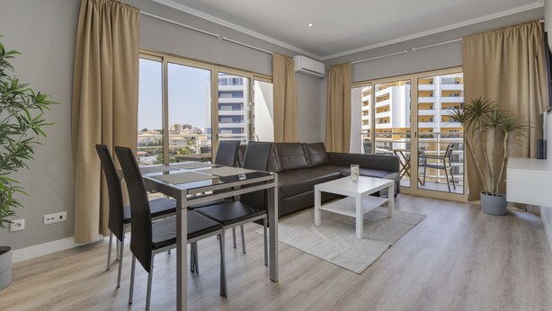 A modern living room with a sofa, dining table, chairs, and large windows offering a city view.