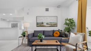 Una sala de estar moderna con un sofá gris, una mesa de centro de madera y un sillón de mimbre. Hay un cuadro en la pared y una planta al lado.