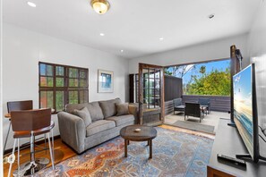Un salón moderno con un sofá, una mesa de centro y un televisor. Hay un ventanal con persianas y una puerta que da a un patio exterior.