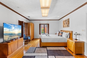 Un dormitorio con una televisión grande, muebles de madera, una cama con almohadas, una ventana y un cuadro en la pared.