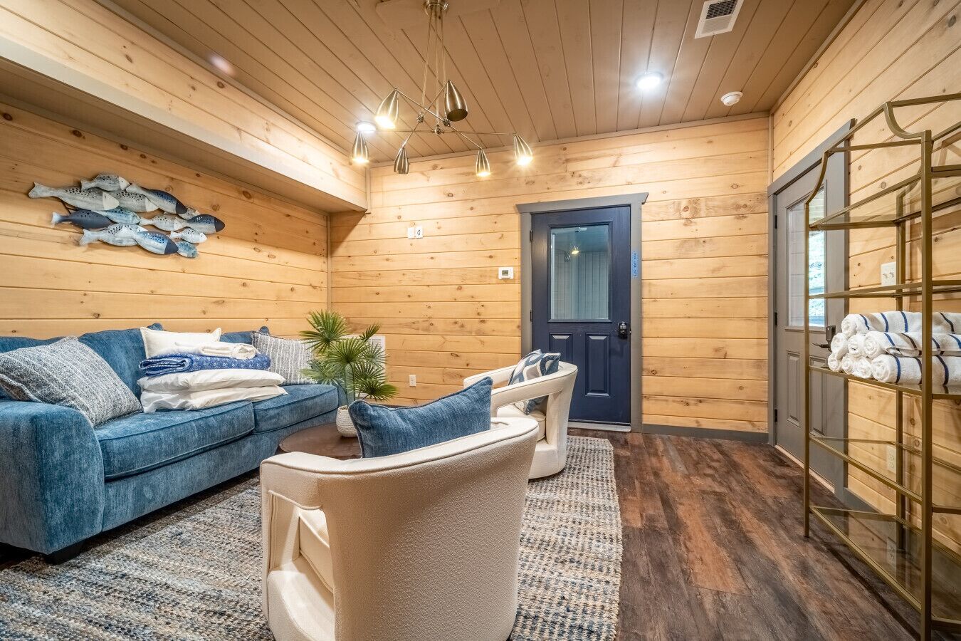 A cozy room with a blue sofa, a beige armchair, and a small table with a plant. There is a wooden door and a shelf with towels.