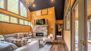 A cozy cabin interior with a stone fireplace, wooden walls, and large windows. - Multi-Level Getaway | Pool, Hot Tub and Fireplace (Sevierville)