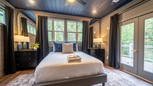 A bedroom with a large bed, dark wooden headboard, two bedside tables with lamps, and a view of greenery through large windows. - Multi-Level Getaway | Pool, Hot Tub and Fireplace (Sevierville)