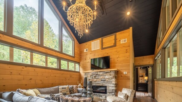 A cozy log cabin interior with a stone fireplace, large windows, and a chandelier. - Multi-Level Getaway | Pool, Hot Tub and Fireplace (Sevierville)