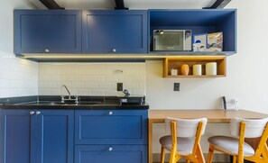 Eine moderne Küche mit blauen Schränken, einer schwarzen Arbeitsplatte und einer Mikrowelle. Zwei Holzstühle und ein kleines Holzregal mit Gläsern sind ebenfalls vorhanden.