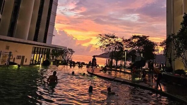 Zona de piscina con gente disfrutando del agua, rodeada de edificios altos y un cielo al atardecer.