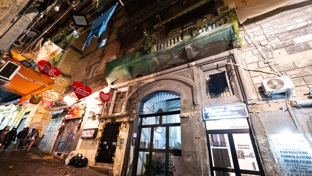 Vista de una calle con un edificio que presenta una entrada arqueada grande, un letrero de una agencia funeraria y una unidad de aire acondicionado visible.