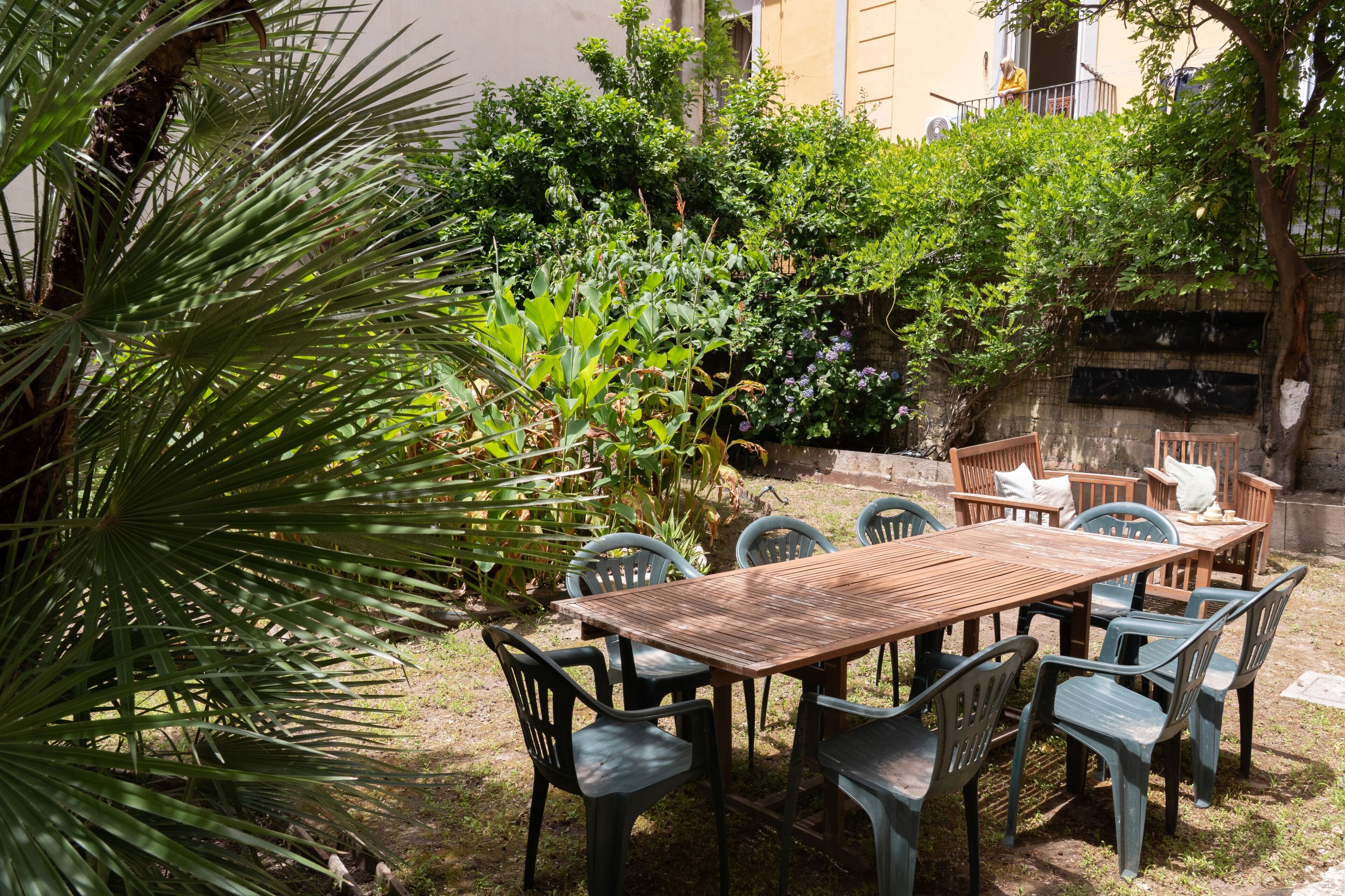 A wooden table with chairs in a garden area with greenery and a brick wall.