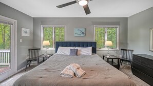 Un dormitorio con una cama grande, dos mesitas de noche, un ventilador de techo y vistas a árboles por las ventanas.