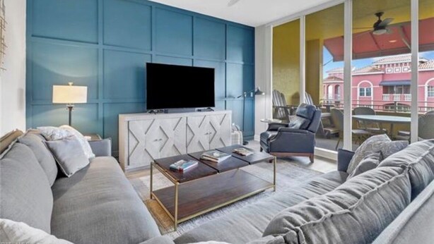 A modern living room with a large flat-screen TV, grey sofas, a coffee table, and a view of a red building through large windows. - Luxury 2 Bed 2 Bath  Penthouse Bayfront Condo off 5th Avenue (Naples)