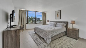 A modern hotel room with a large bed, a desk, a TV, and a view of the city. - Hallandale Beach - 2 BR Luxury Condo (Hollywood)
