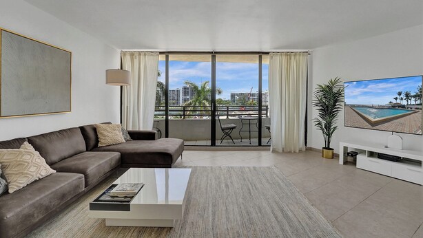 A modern living room with a sofa, a coffee table, a TV, and a balcony with a view of the city. - Hallandale Beach - 2 BR Luxury Condo (Hollywood)