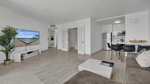 A modern living room with a flat-screen TV, a coffee table, and a sofa. There is a kitchen area with a refrigerator, stove, and bar stools. - Hallandale Beach - 2 BR Luxury Condo (Hollywood)