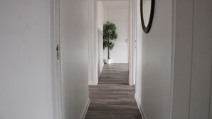 A hallway with white walls and wooden flooring, leading to a room with a potted plant.