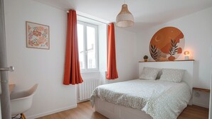 A bedroom with a bed, a window with curtains, a bedside table with a lamp, and a wall art piece. - Charming T2 in the center of Saint-Astier (Saint-Astier)
